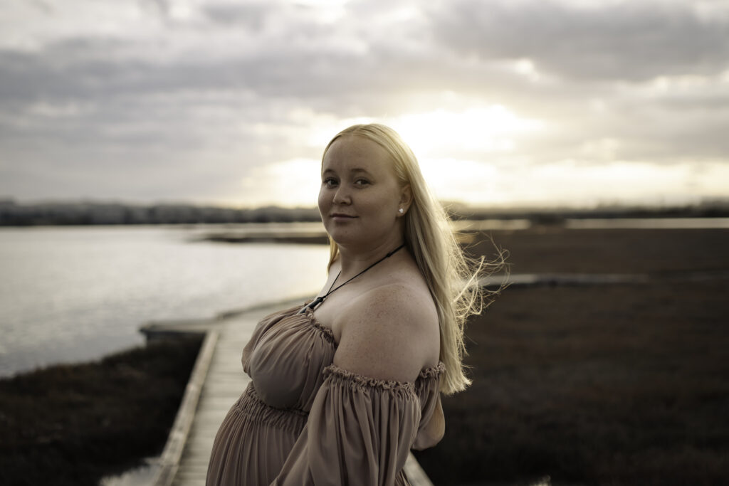 Maternity session at Pandora Estuary, Hawke’s Bay, at sunset. First-time mother standing in shallow water, wearing a flowing dress, soft golden light reflecting on the estuary.