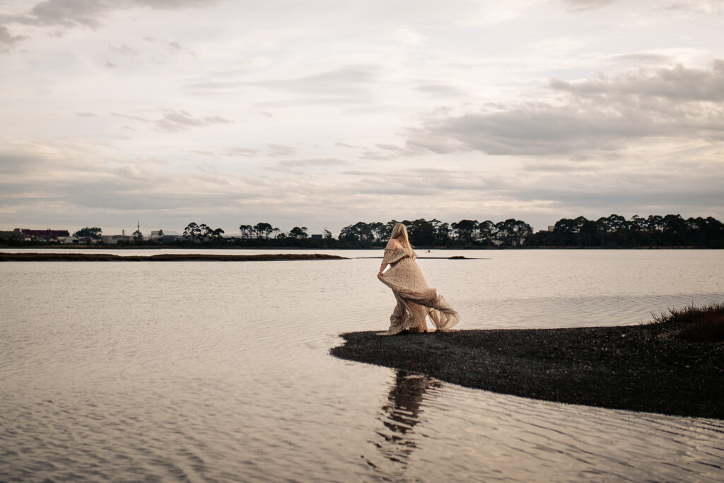Maternity photography at Pandora Estuary, Hawke’s Bay. Woman swishing flowing dress on a small island in the water at sunset.