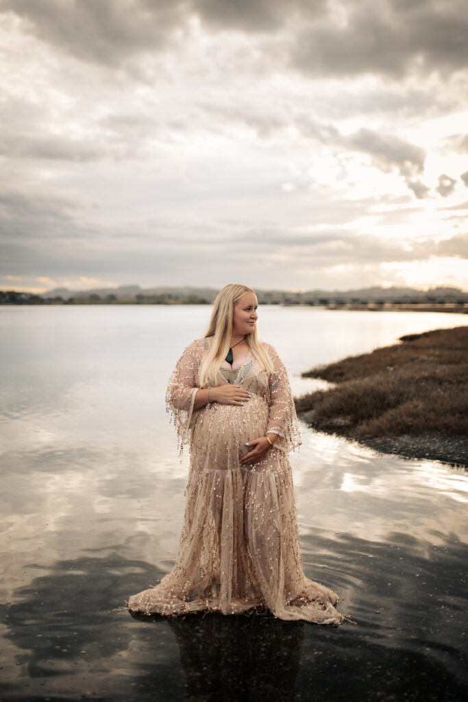 Maternity photography at Pandora Estuary, Hawke’s Bay. Woman standing in shallow water at sunset with reflections on the estuary surface.