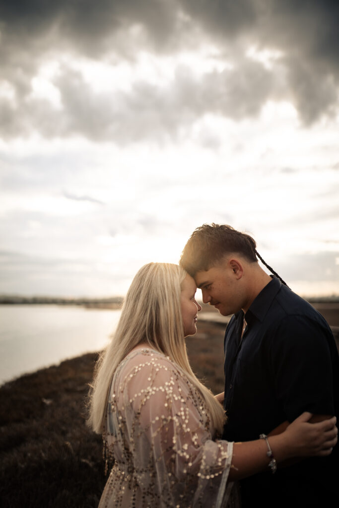 Pregnant couple at Pandora Estuary, Hawke’s Bay, foreheads together with sun flare between them during sunset maternity photography.