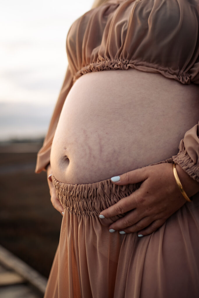Pregnant woman at Pandora Estuary, Hawke’s Bay holding her bare belly during a sunset maternity session.
