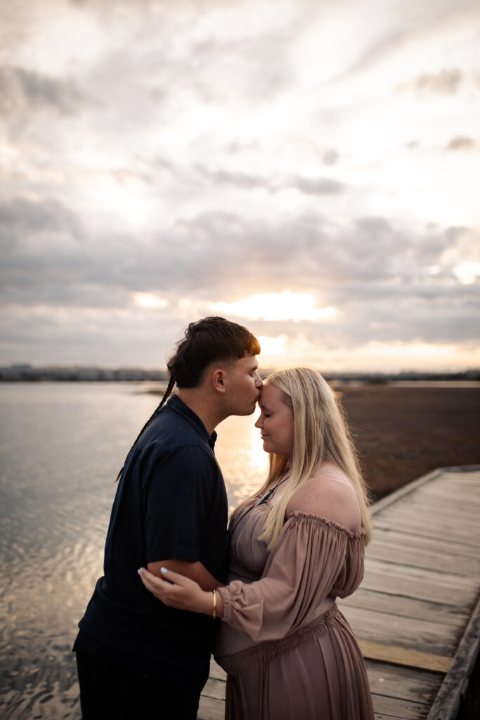 Pregnant woman and partner at Pandora Estuary, Hawke’s Bay. Partner kisses her forehead during a sunset maternity session.